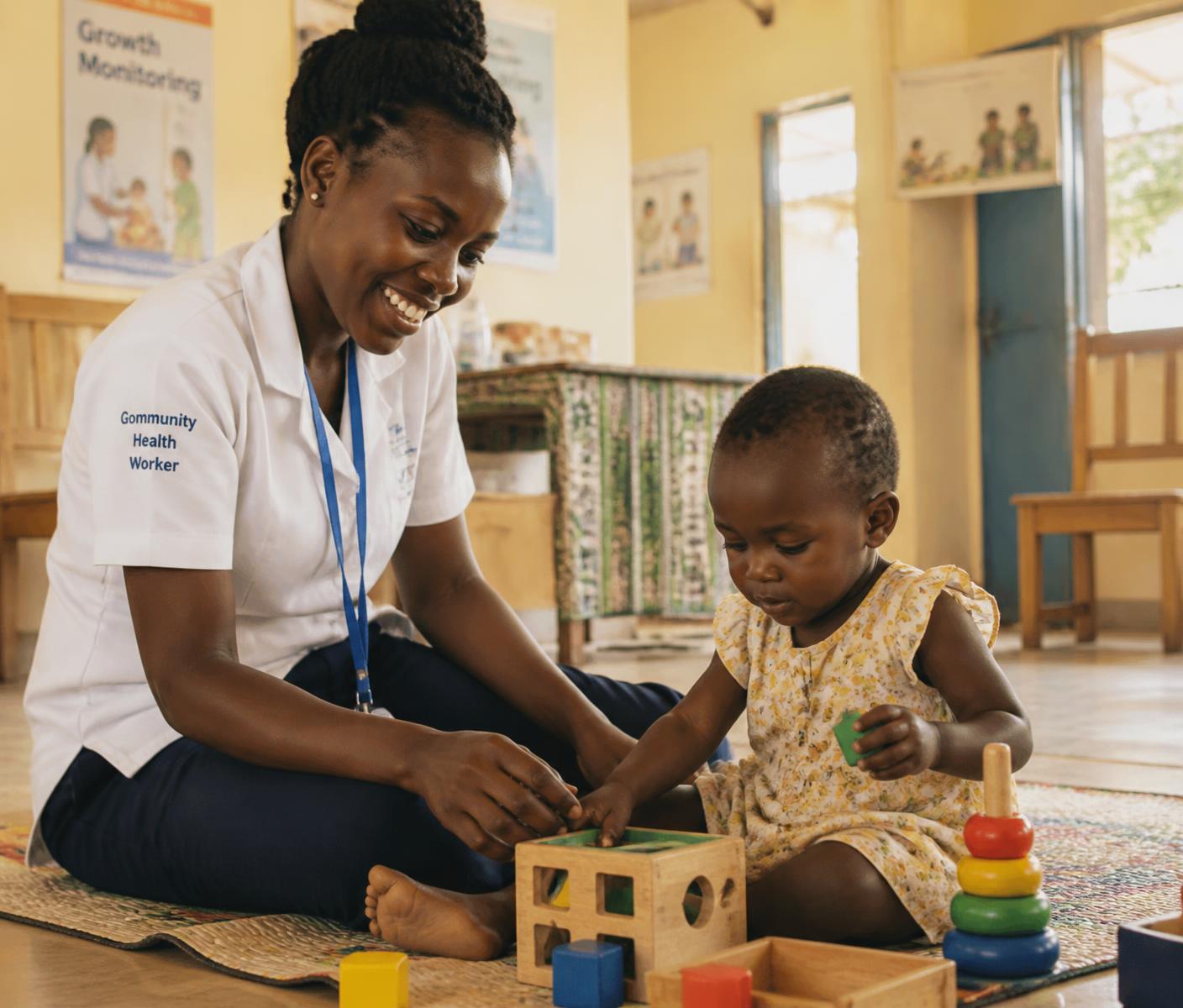 A community health worker sitting on the floor beside a toddler playing with developmental toys in an African clinic